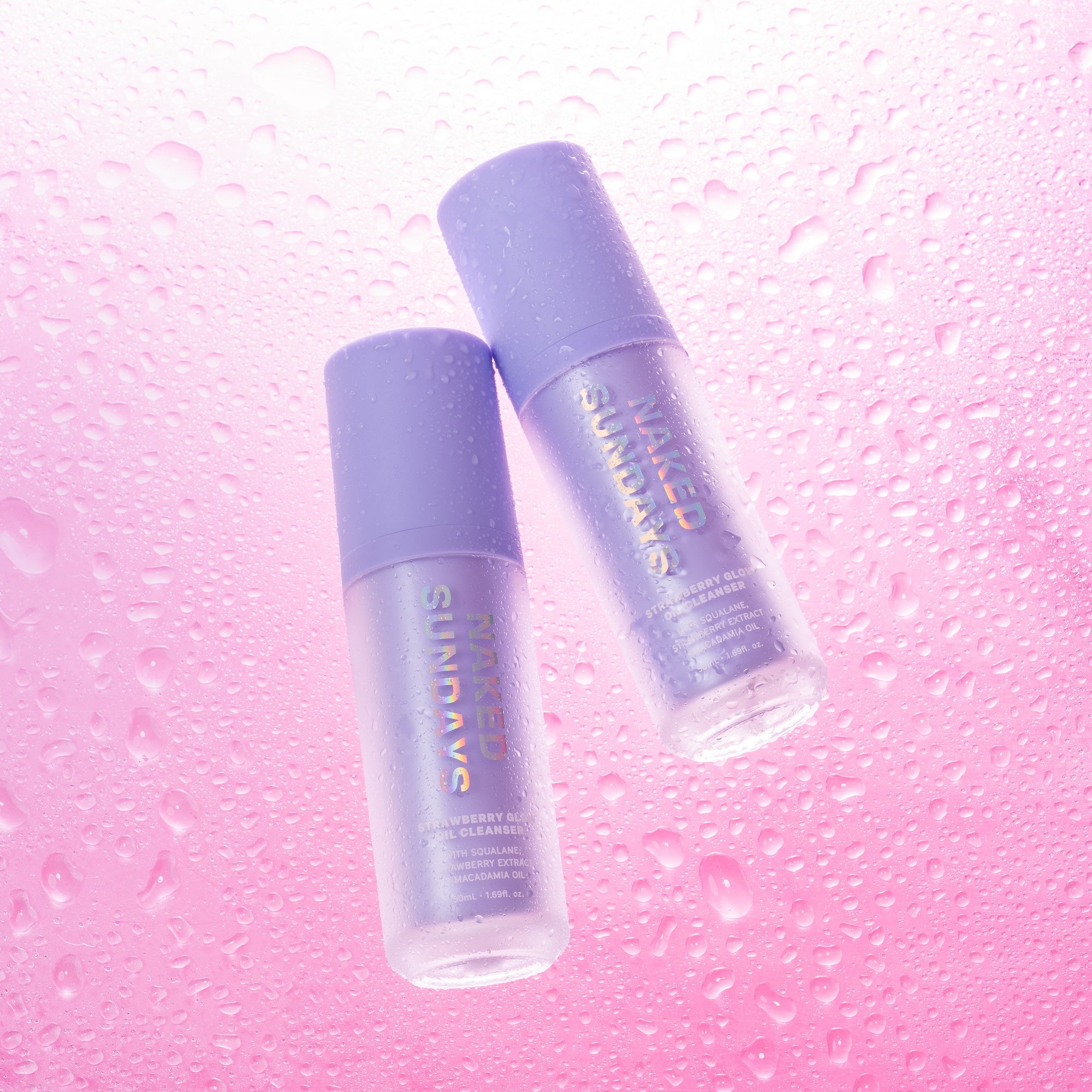 Two bottles of Strawberry Glow Oil Cleanser against a pink background with water droplets.