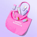 Pink tote bag with 'Naked Sundays' text containing beauty products on a light purple background