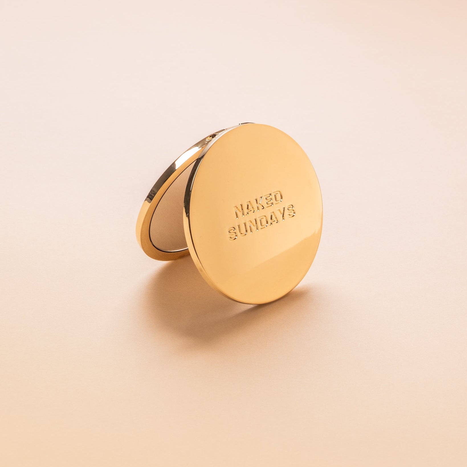 Gold Compact mirror with 'Naked Sundays' branding on a beige background