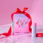 Naked Sundays Rosy Glow Kit beauty gift set featuring a pink box tied with a red ribbon, a lip product tube, a face product tube, and a spray bottle, arranged on a pink glittery surface against a pink background.