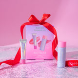 Naked Sundays Rosy Glow Kit beauty gift set featuring a pink box tied with a red ribbon, a lip product tube, a face product tube, and a spray bottle, arranged on a pink glittery surface against a pink background.