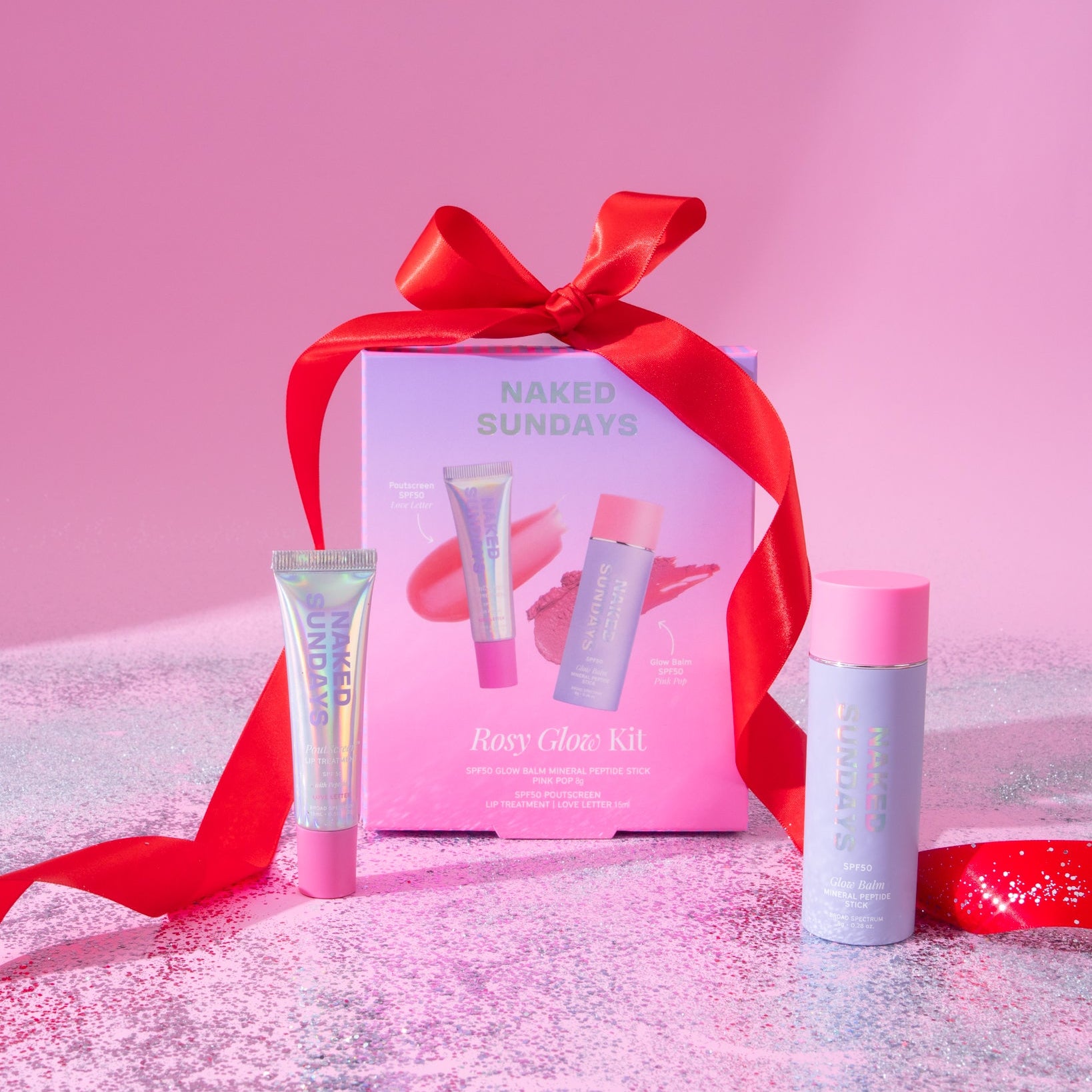 Naked Sundays Rosy Glow Kit beauty gift set featuring a pink box tied with a red ribbon, a lip product tube, a face product tube, and a spray bottle, arranged on a pink glittery surface against a pink background.