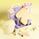 A yellow travel-themed beauty gift set arranged in an open suitcase on a pale yellow background. The set includes a cream cap with 'Naked Sundays' text, a yellow pouch, purple and pink cosmetic tubes, a small mirror, and a colorful patterned card.