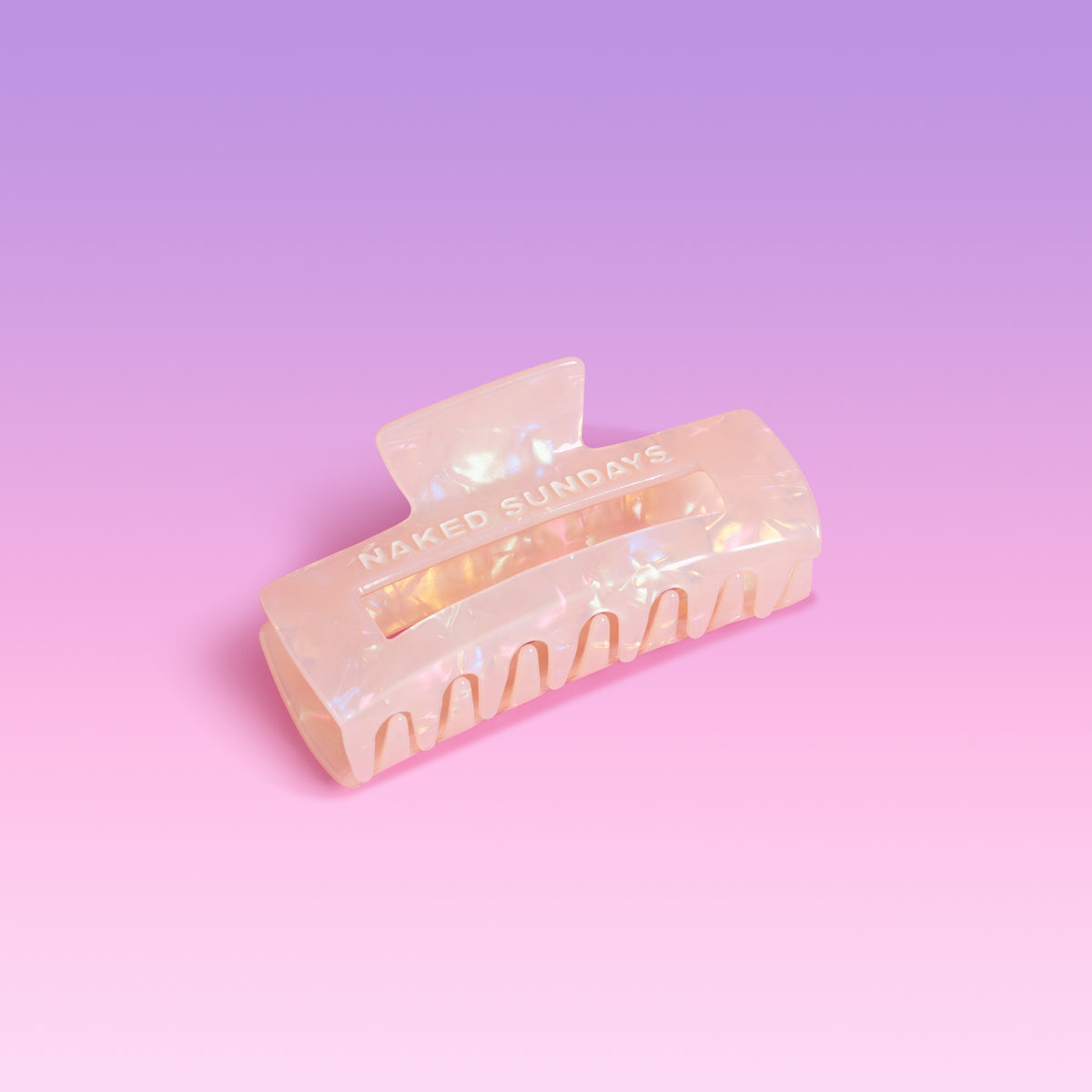 A translucent yellow-colored claw hair clip with an iridescent finish, featuring the text 'NAKED SUNDAYS' printed on the side, set against a soft pink and purple gradient background.
