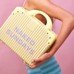 A woman's arm is shown cradling a yellow ribbed suitcase with the purple text NAKED SUNDAYS on the front.