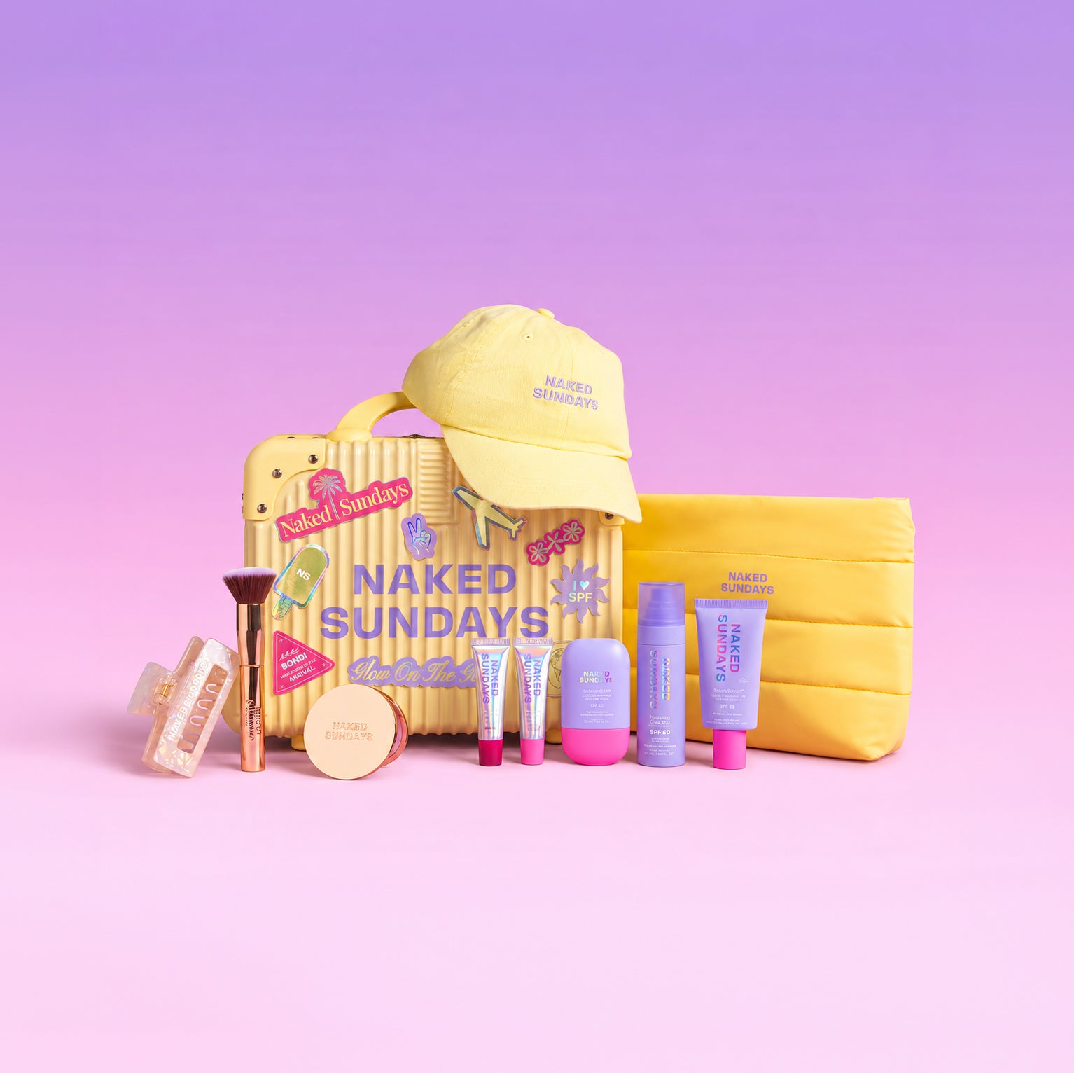 A curated beauty gift set arranged on a pastel pink-to-purple gradient background. The set includes a yellow mini suitcase with 'NAKED SUNDAYS' text, a yellow cap, a yellow makeup pouch, several purple and lavender cosmetic tubes, a brush, and small makeup items.