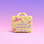 A small yellow ribbed suitcase with a handle, decorated with colorful travel-style stickers and the words 'NAKED SUNDAYS' on the front, set against a soft pink-to-purple gradient background.